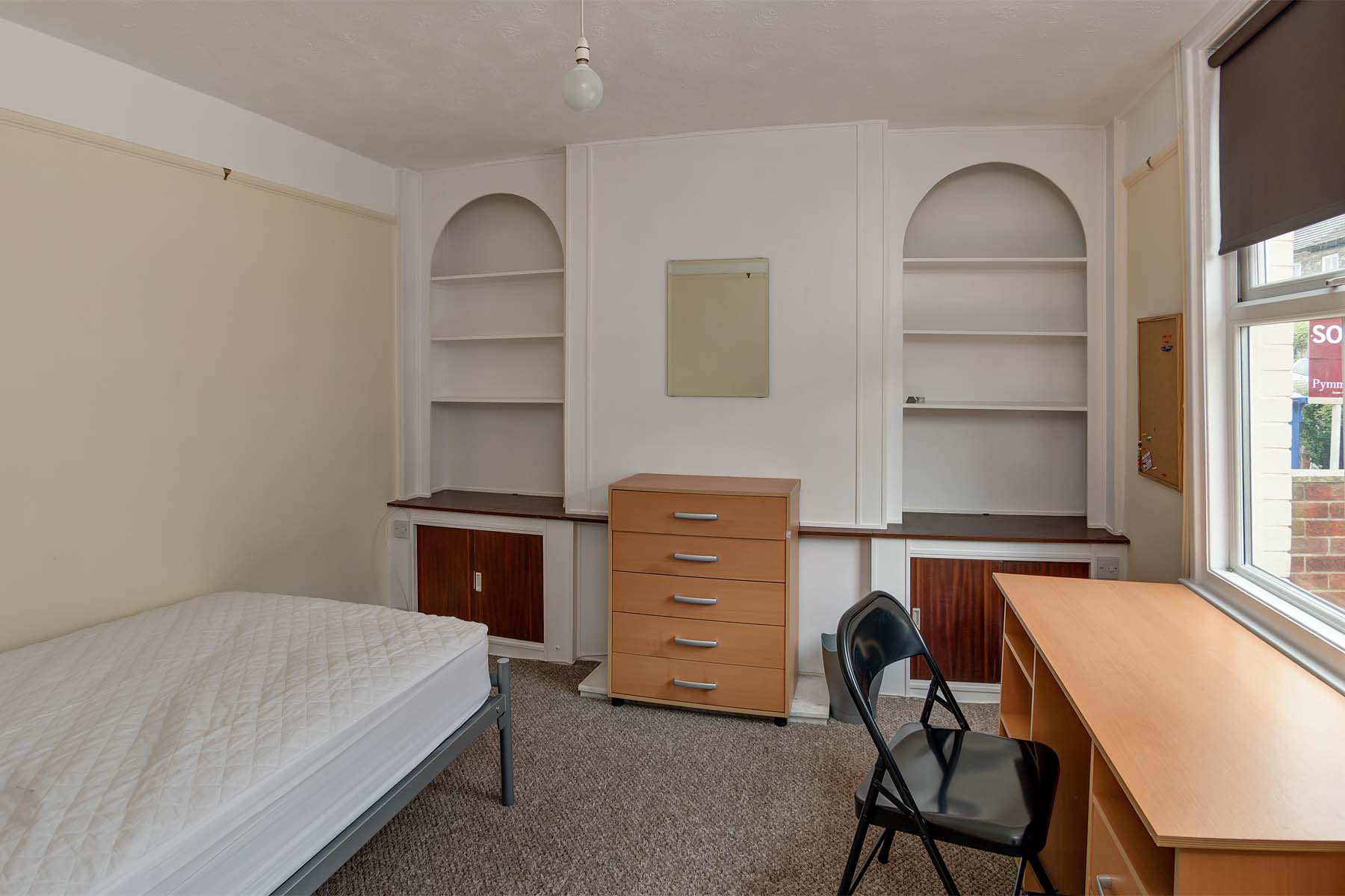 Bedroom with built in arch shaped shelving, with wooden chest of drawers, wooden desk with a black chair and a metal bedframe.