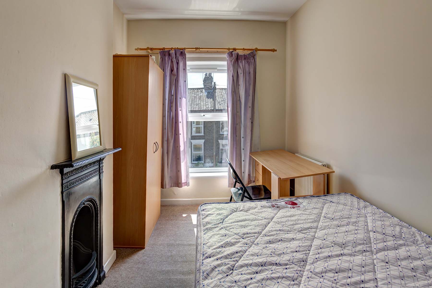 Double bedroom with wooden desk and chair, a wooden wardrobe and a metal bedframe and mattress. The room also features a decorative fireplace.