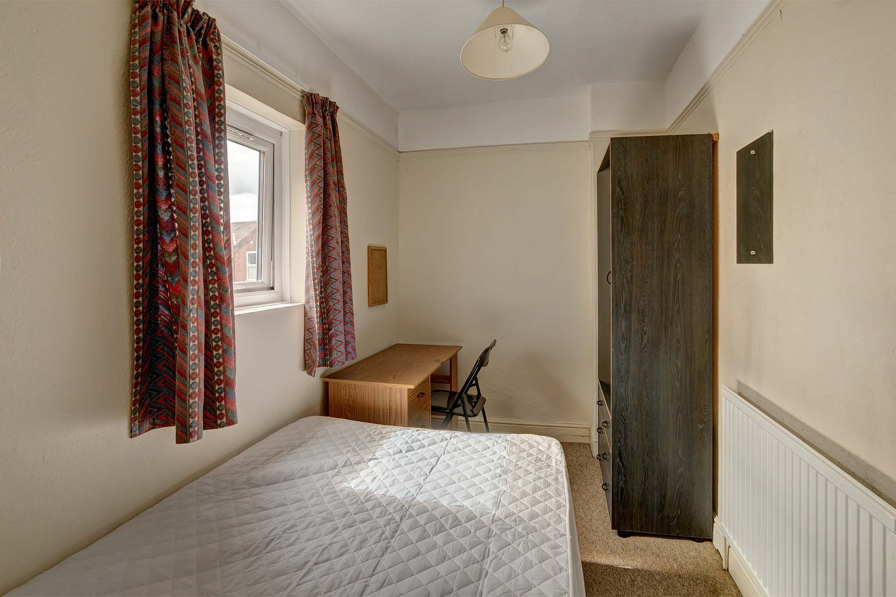 Double bedroom with bed base and mattress, a dark grey wood effect wardrobe and a wooden desk and black chair.