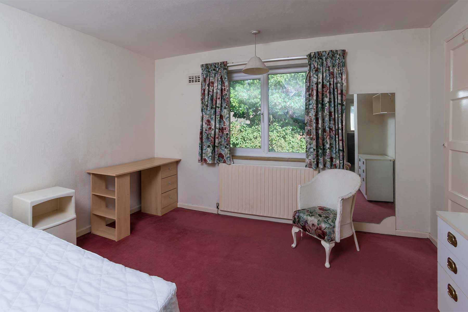 Bedroom with a red carpet and pattered curtains. A wooden desk, bedside table and chest of drawers.