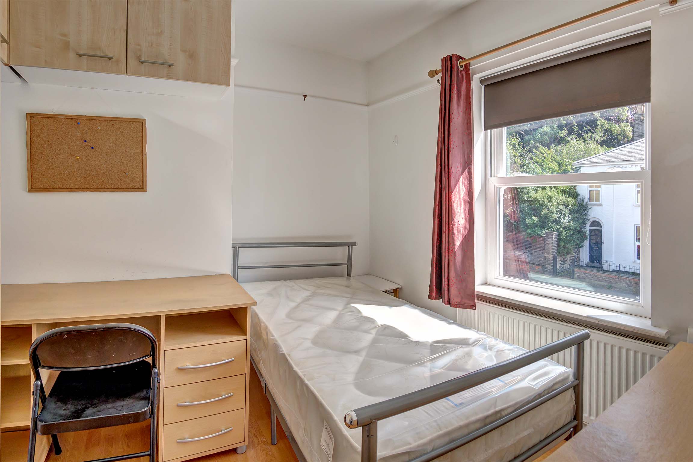 Bedroom with metal bed frame, wooden desk and chair and cupboard space fitted to the wall.
