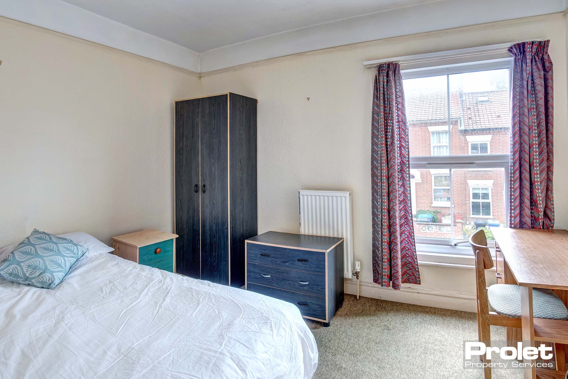 Double bedroom with dark grey wooden wardrobe, blue chest if drawers and a bedside table. Wooden desk and chair.