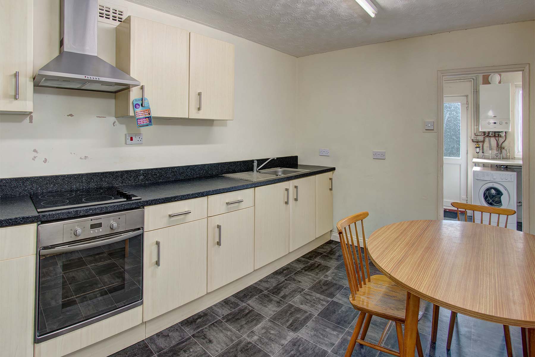 White fitted kitchen with dark grey tiled flooring and a wooden dining table with chairs.