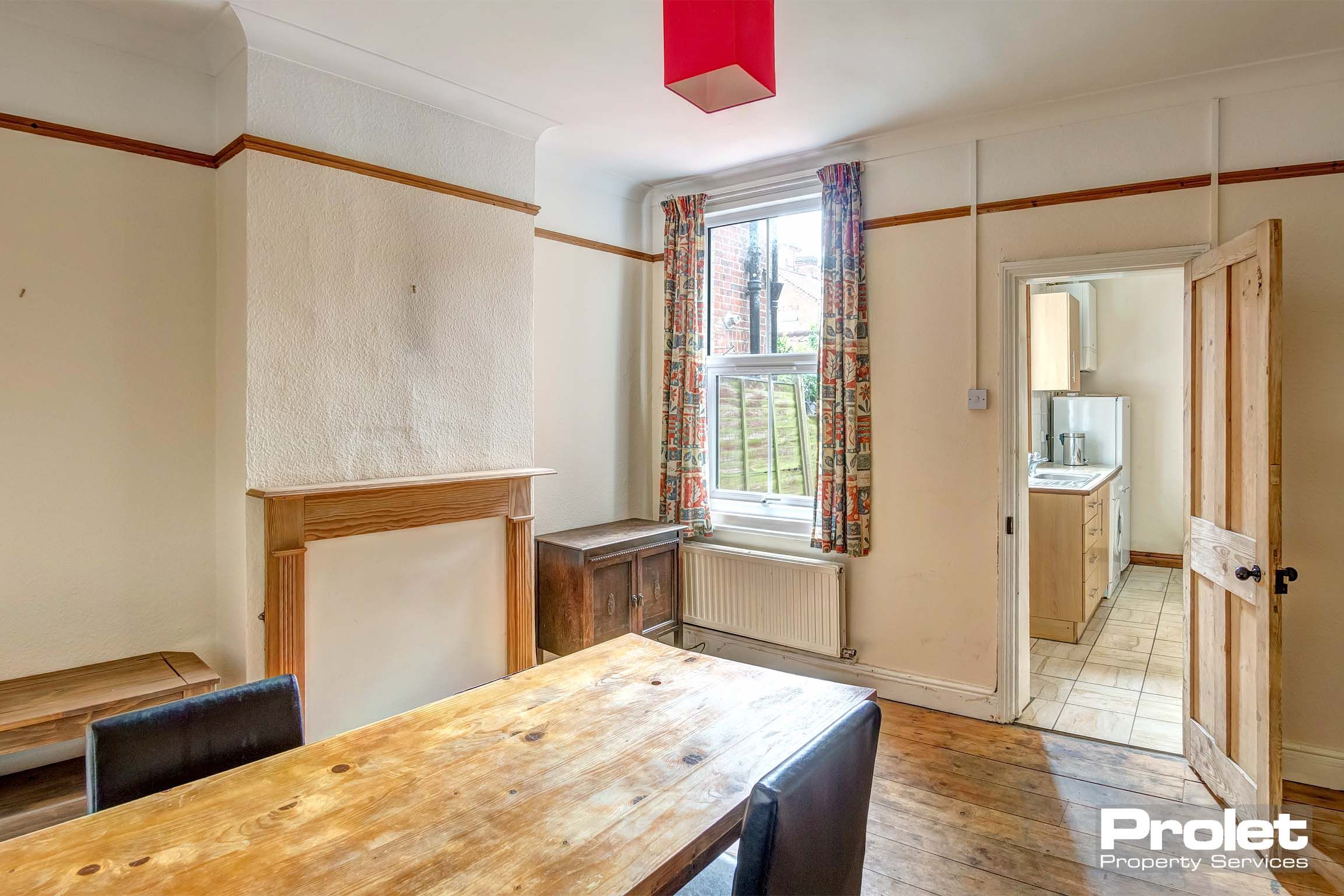 Dining room with wooden dining table and chairs. Decorative wooden fireplace. Red lampshade and patterned curtains.