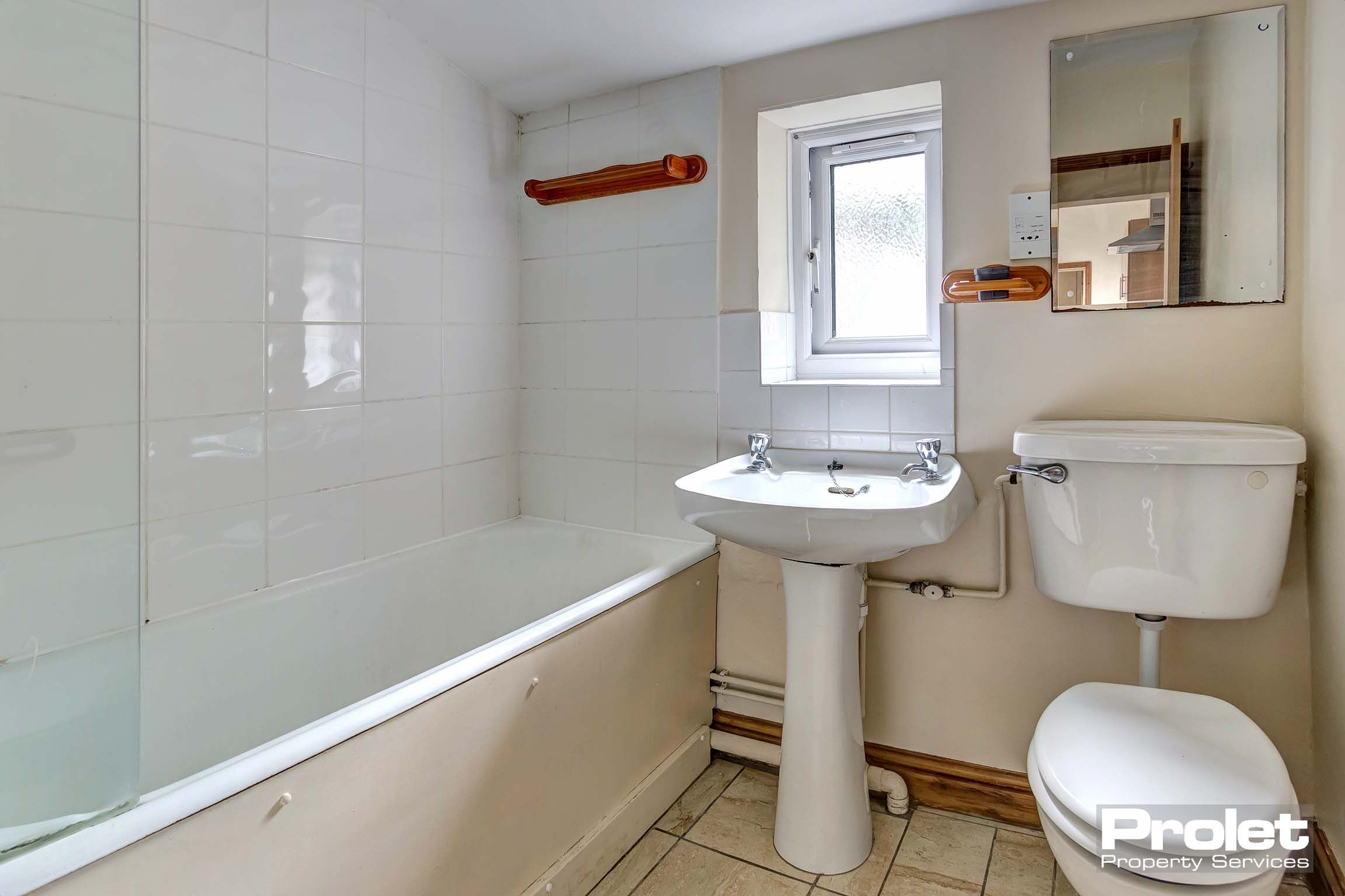 White tiled bathroom with a white sink, toilet and bath with a shower over the bath and a glass splash guard.