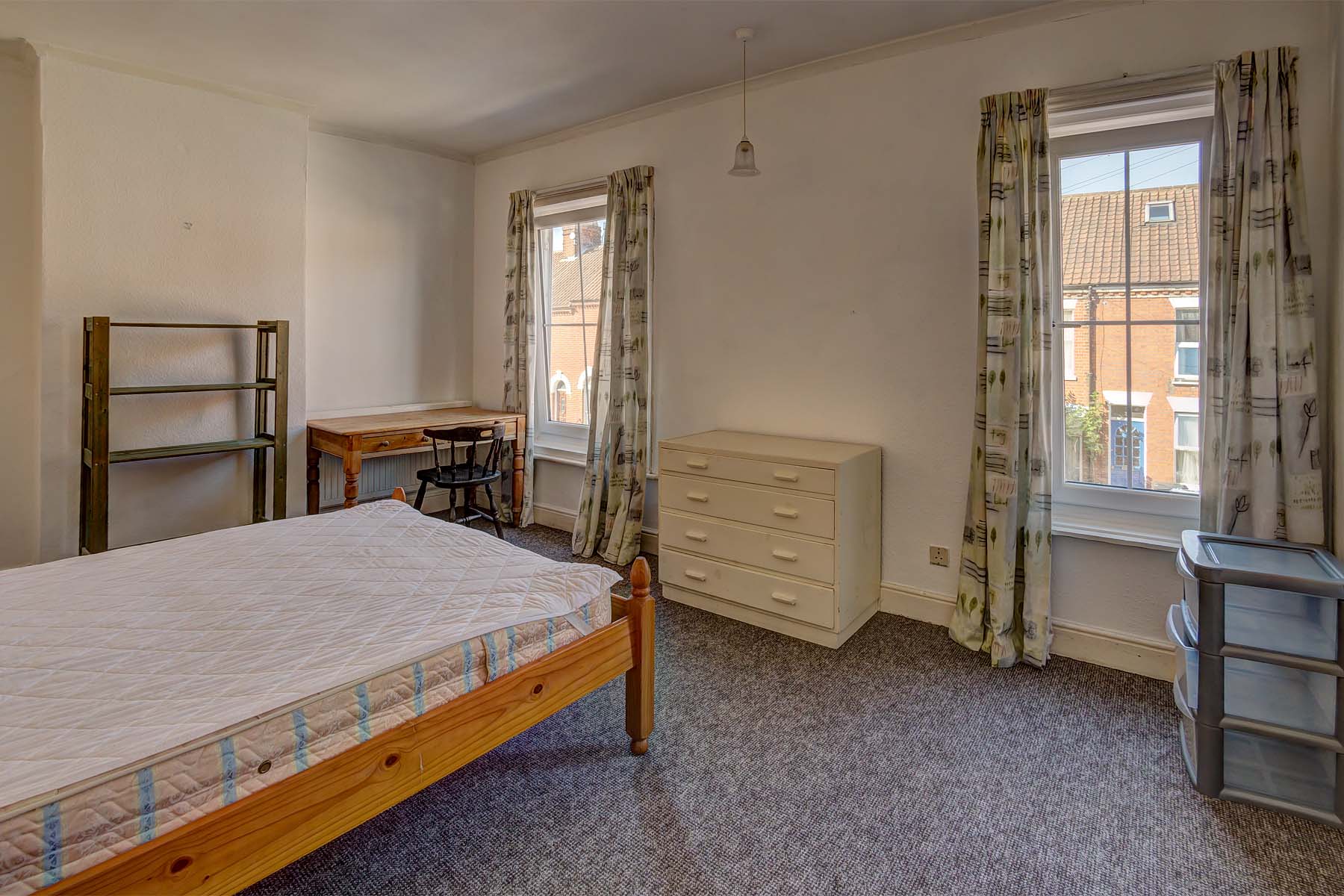 Large double bedroom with two windows, a wooden bed frame and mattress. A white set of drawers and wooden desk and chair.