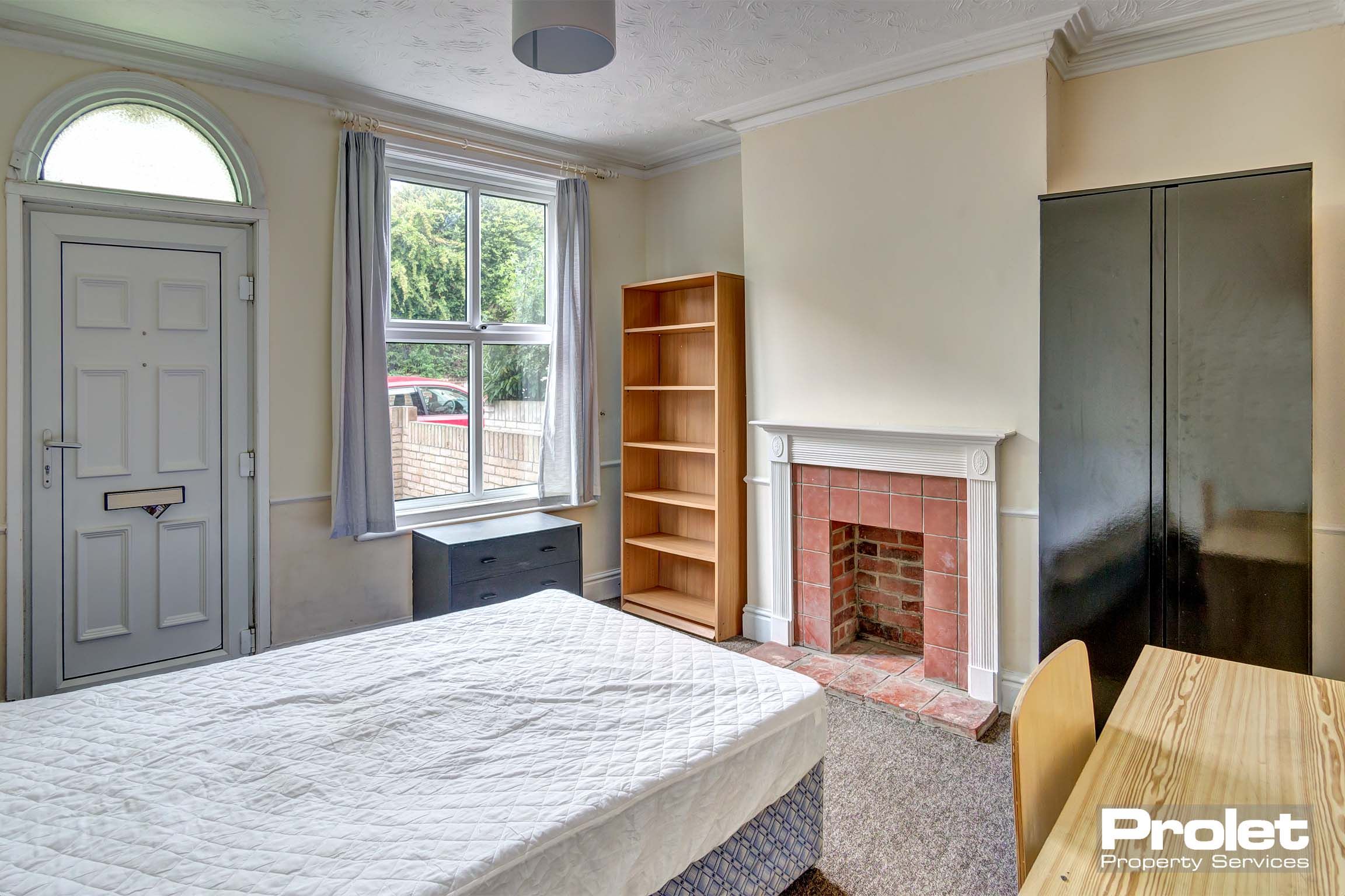 Double bedroom with front door entry/exit. A divan bed base and mattress, a black wardrobe, a wooden desk and bookcase and a decorative brick fireplace.
