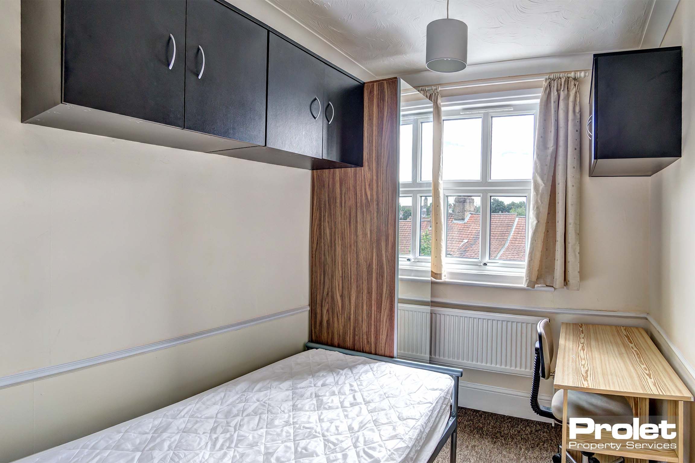 Bedroom with metal bed frame and mattress. Wooden desk and chair. Black storage cupboards fixed to walls.