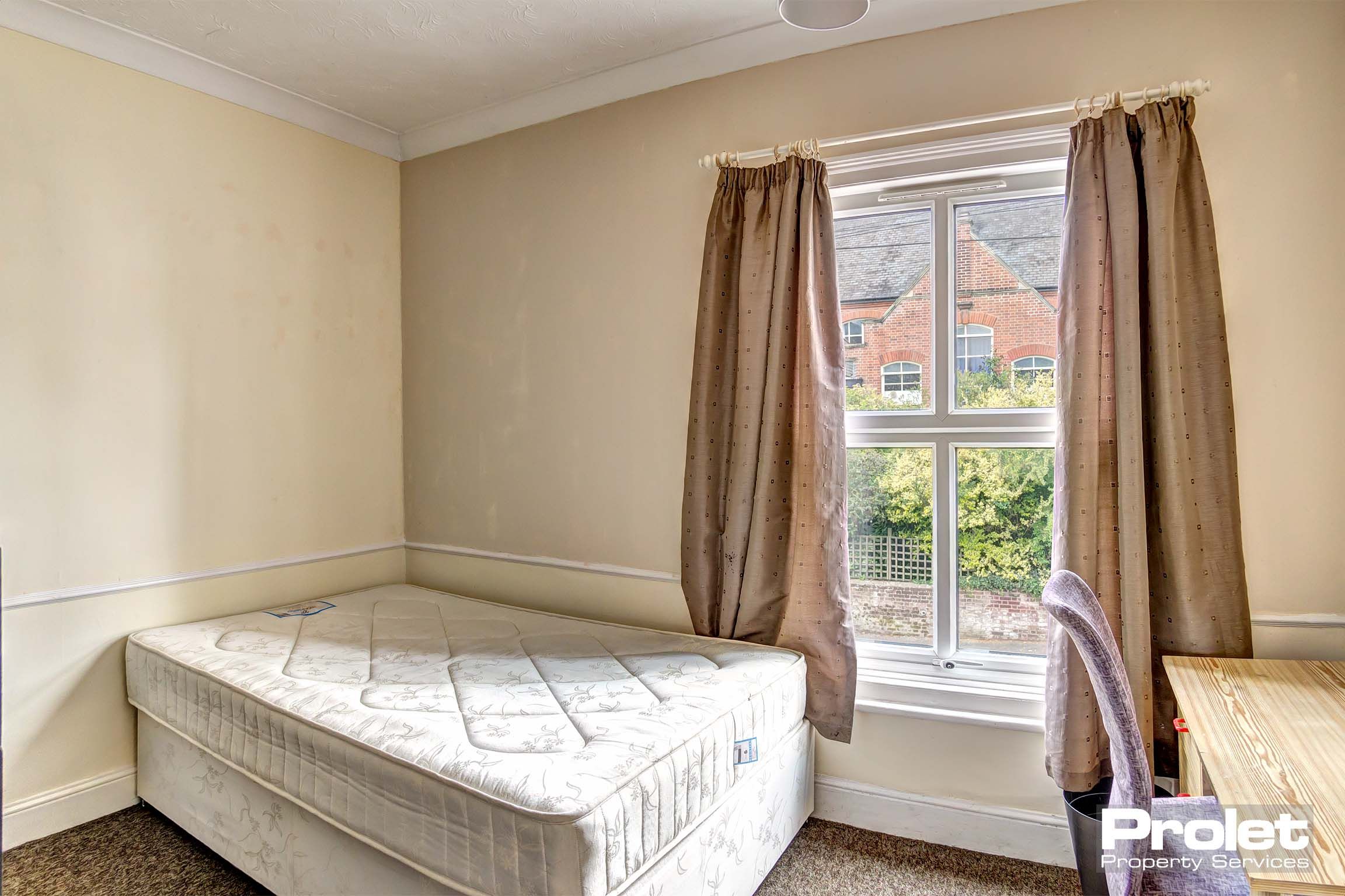Double bedroom with divan bed base and mattress. A wooden desk and chair. Light brown carpet and curtains.