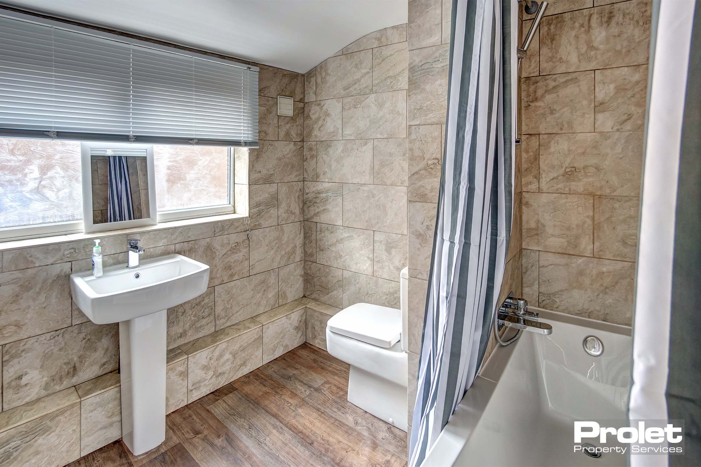 Modern tiled bathroom with shower over bah, sink, and toilet.