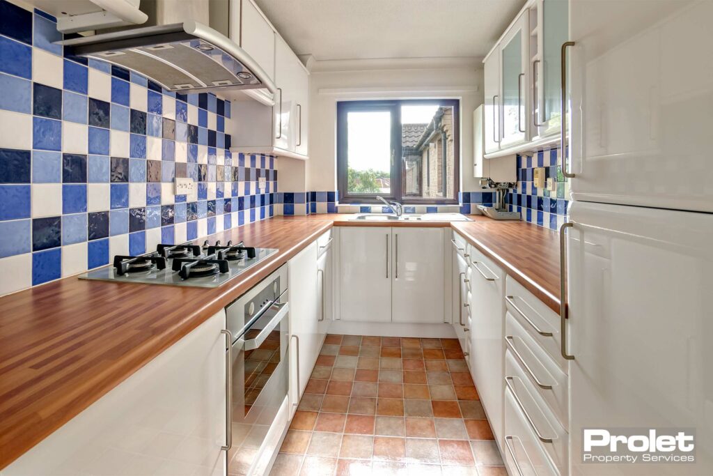Terracotta coloured floor with beech effect work tops, white units and silver handles. Blue mosaic wall tiles and large window over sink. Oven, intergarted washing machine and fridge freezer.