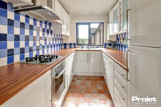 Terracotta coloured floor with beech effect work tops, white units and silver handles. Blue mosaic wall tiles and large window over sink. Oven, intergarted washing machine and fridge freezer.