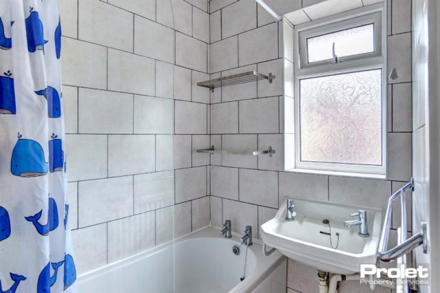 White tiled bathroom with bathtub and shower over the bath. White sink and blue shower curtain.