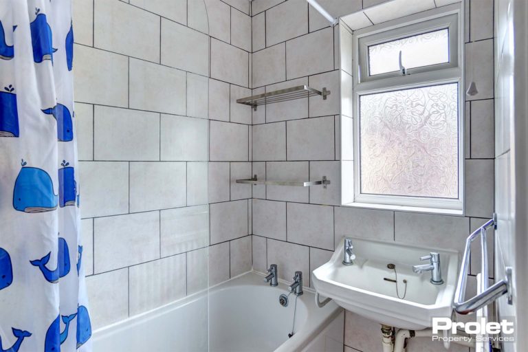 White tiled bathroom with bathtub and shower over the bath. White sink and blue shower curtain.