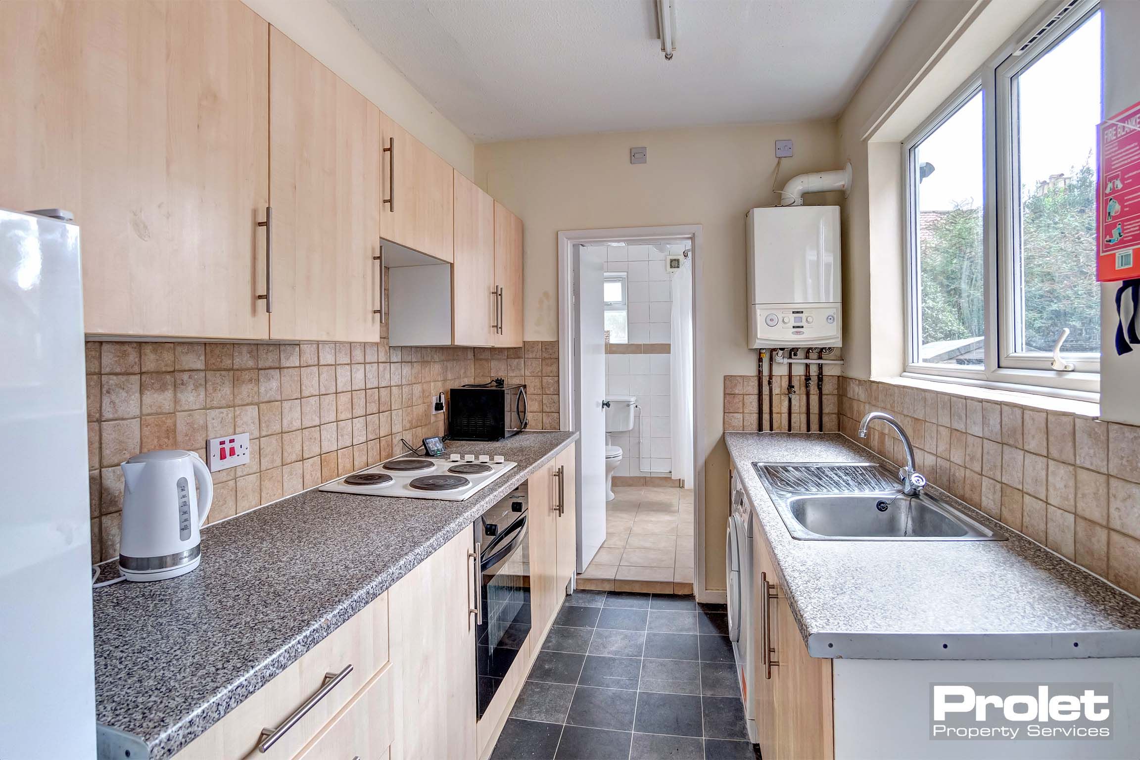 Long galley kitchen with wood effect cabinets, tiled floor, and white goods, leading to bathroom