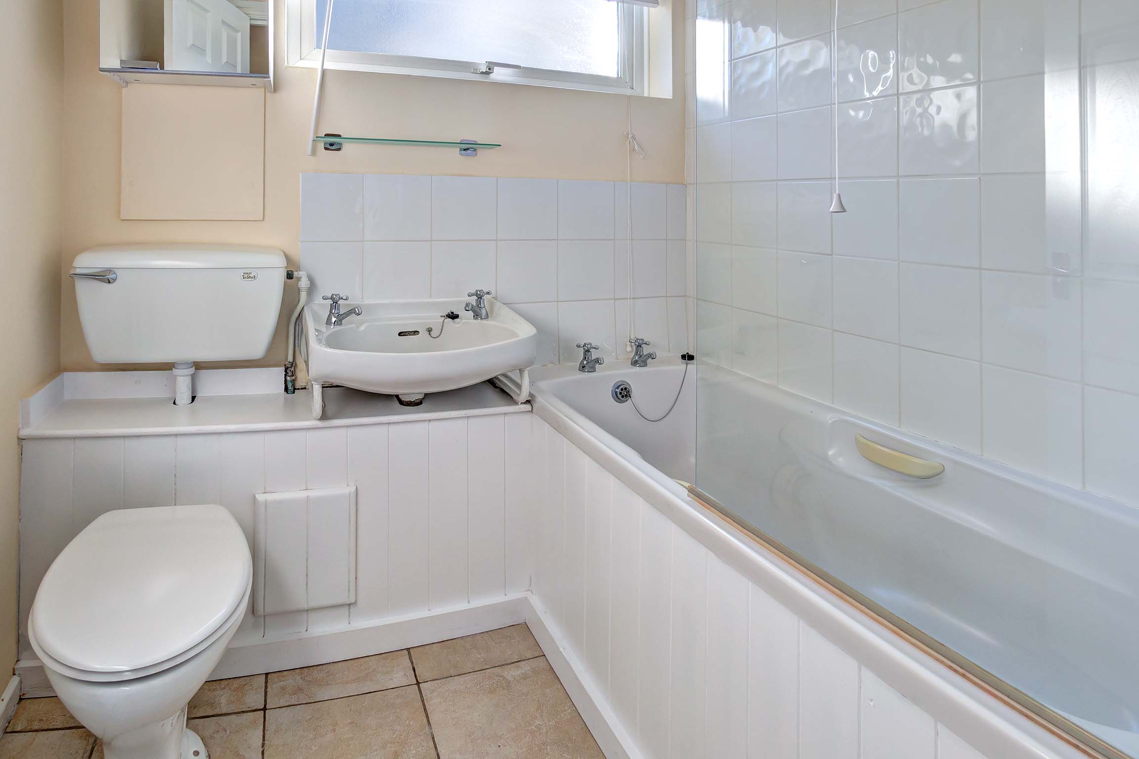 Tiled bathroom with a white toilet and sink. A white bath tub with a glass shower guard and a shower above the bath.