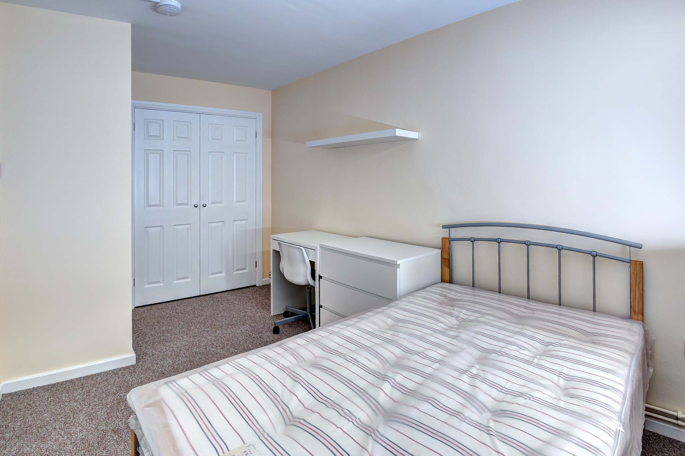 Double bedroom with brown carpet and magnolia walls. A built in white double wardrobe, a white desk with chest of drawers, a white shelf above and a double bed frame and mattress.