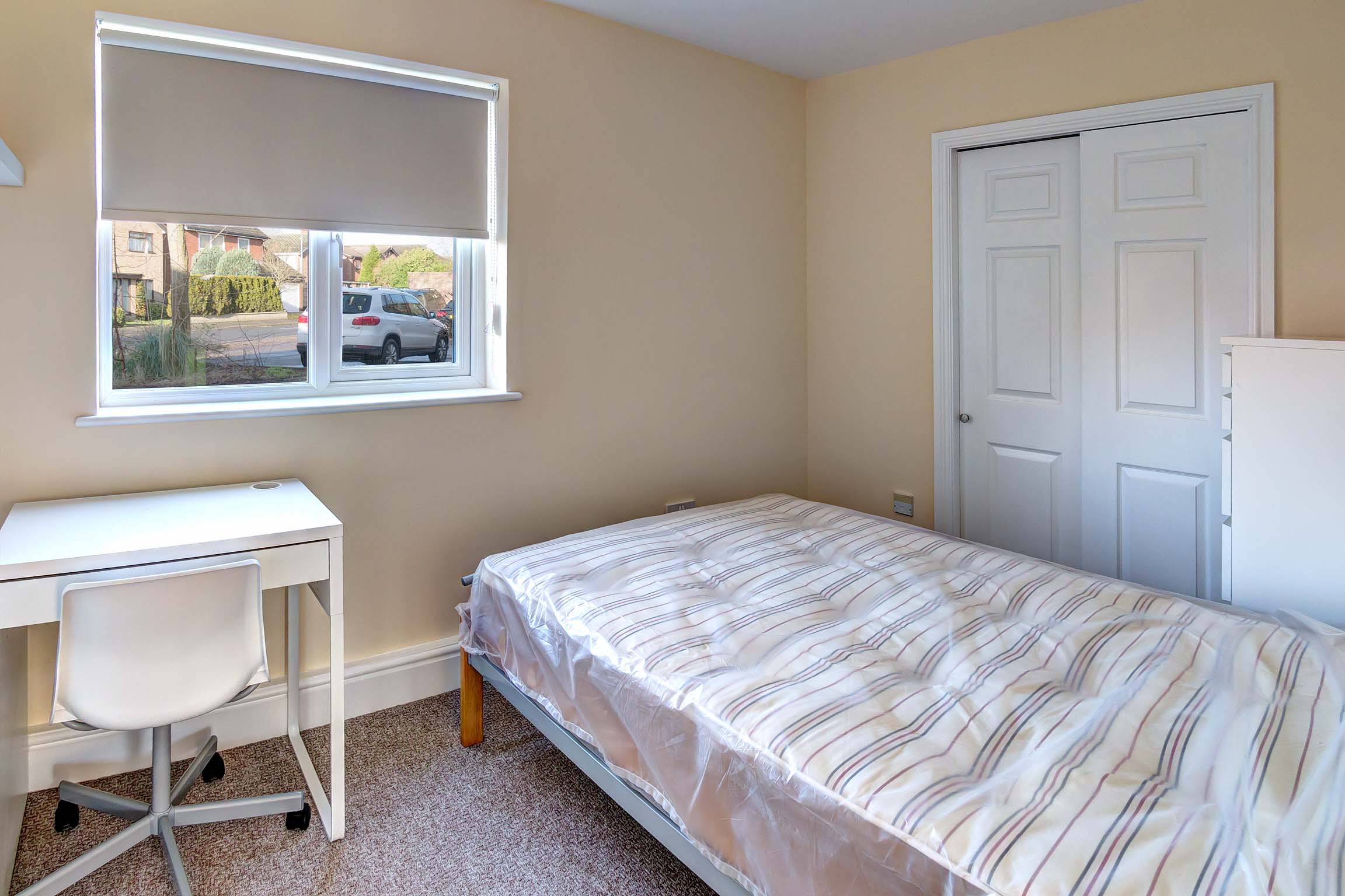 Double bedroom with brown carpet and magnolia walls with a white built in wardrobe, a white desk and chair and a double bed frame with a mattress.