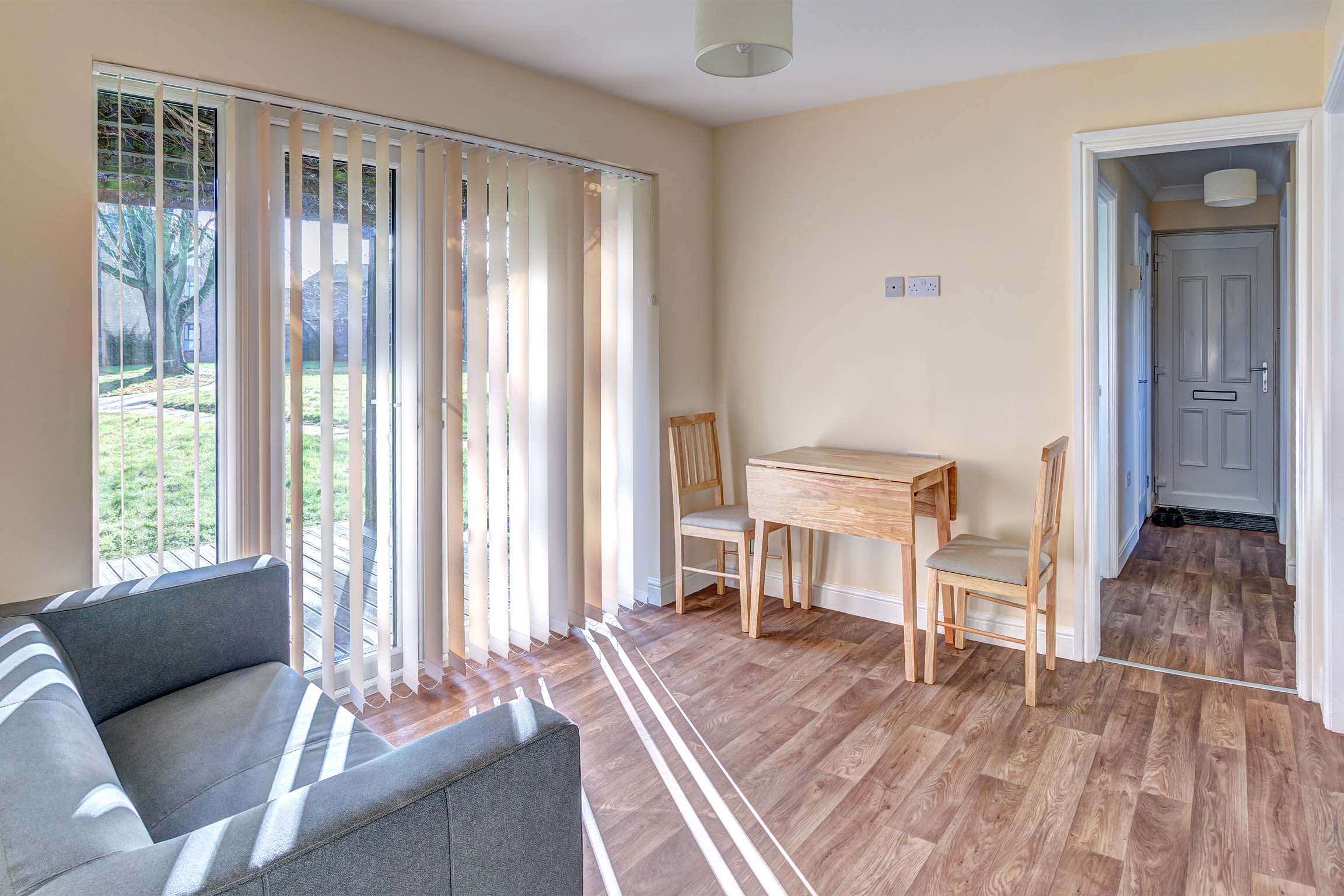 Lounge area with wooden effect lino flooring, a dining table for two and a grey fabric sofa.