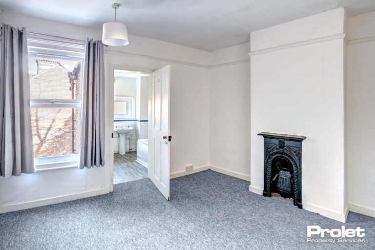 Bedroom with walls painted off white with grey carpet and feature fireplace.