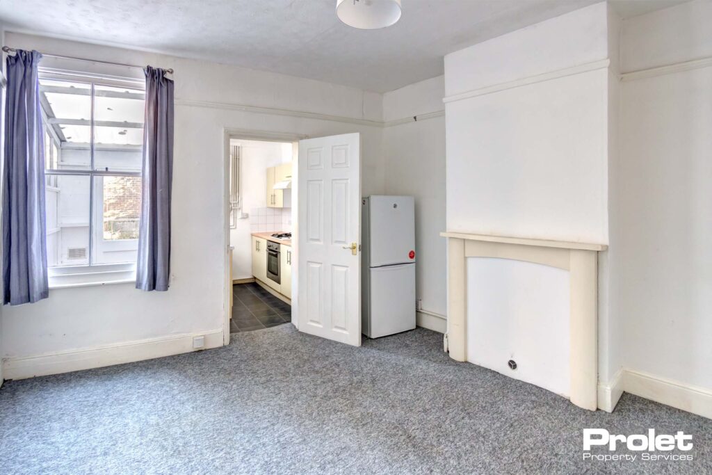 Dining room with walls painted off white with grey carpet and freestanding fridge freezer leading to kitchen