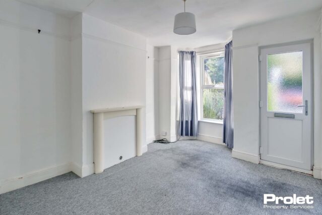 Lounge with bay window. Walls painted off white with grey carpet and boarded up fireplace.