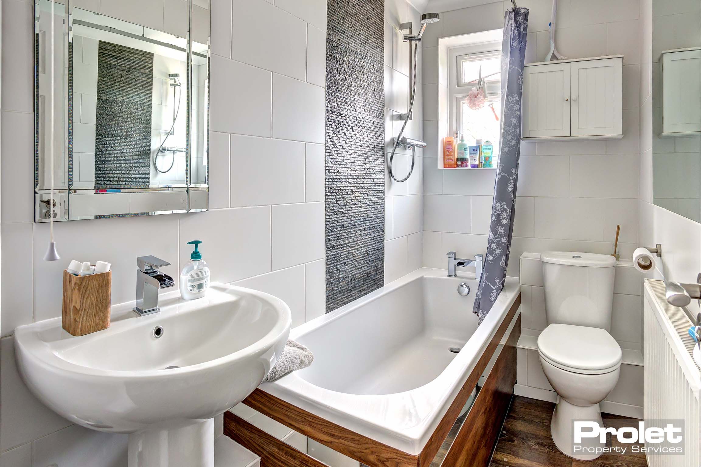 Bathroom with white tiles, a white sink and toilet. A white bath tub with a wooden side panel and a shower over the bath.