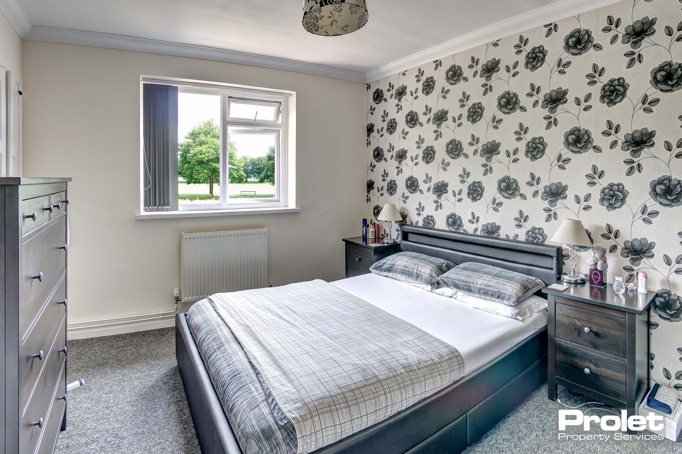 Large double bedroom with flower patterned wallpaper, a double bed frame and matching dark coloured bedside table and chest of drawers