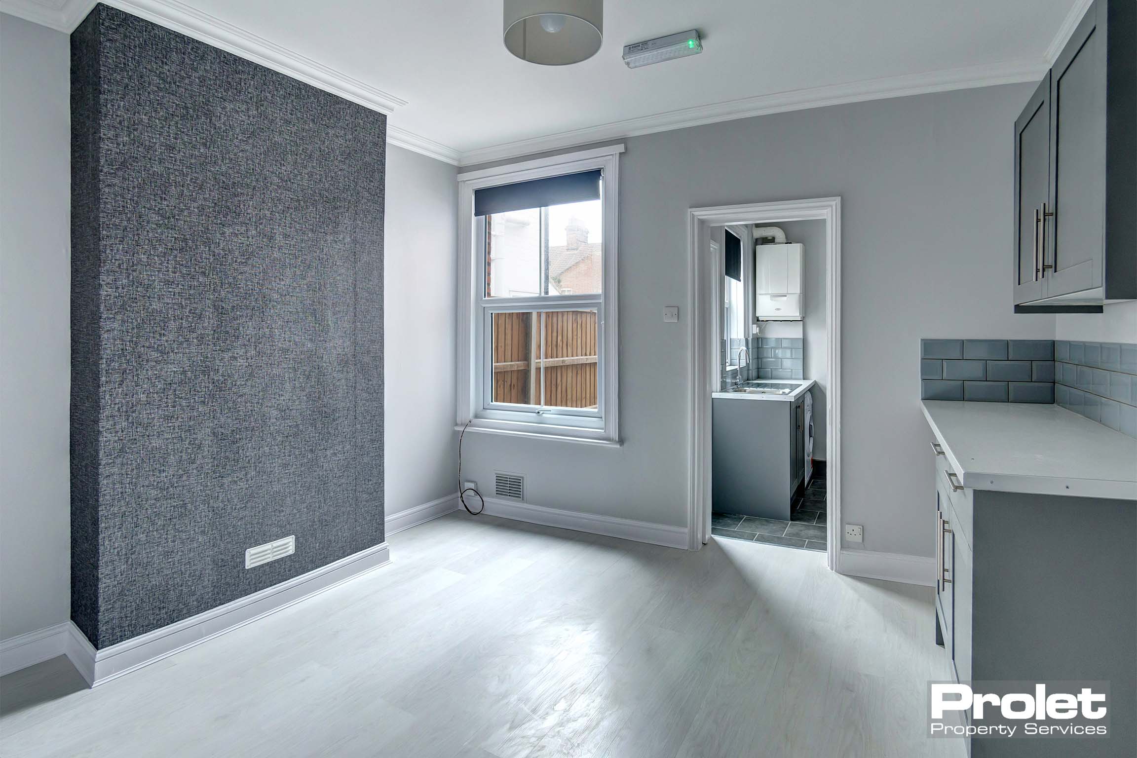 Grey walls and grey vinyl to dining room leading to kitchen. Large window with roller blind