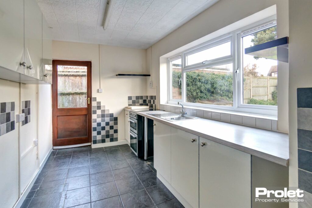 Kitchen with white units and grey tiled floor. Large window over looking rear garden with wooden back door