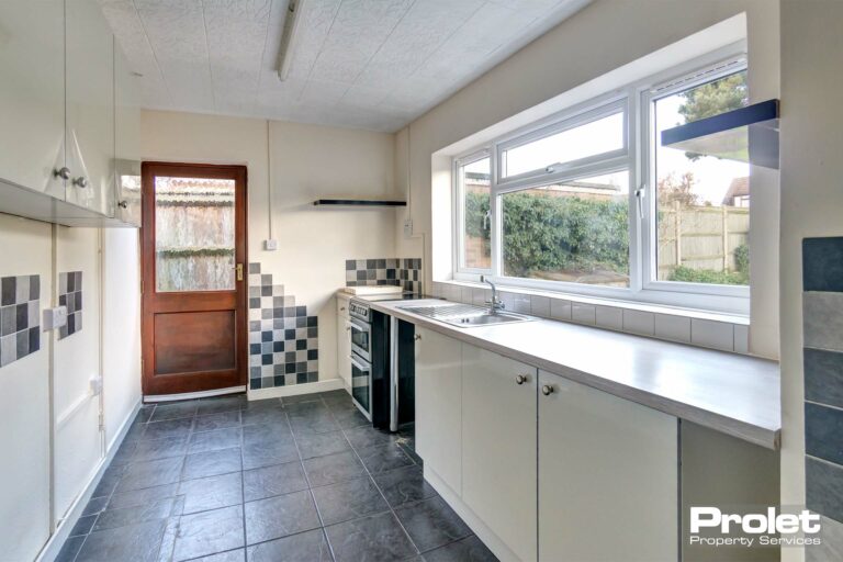 Kitchen with white units and grey tiled floor. Large window over looking rear garden with wooden back door