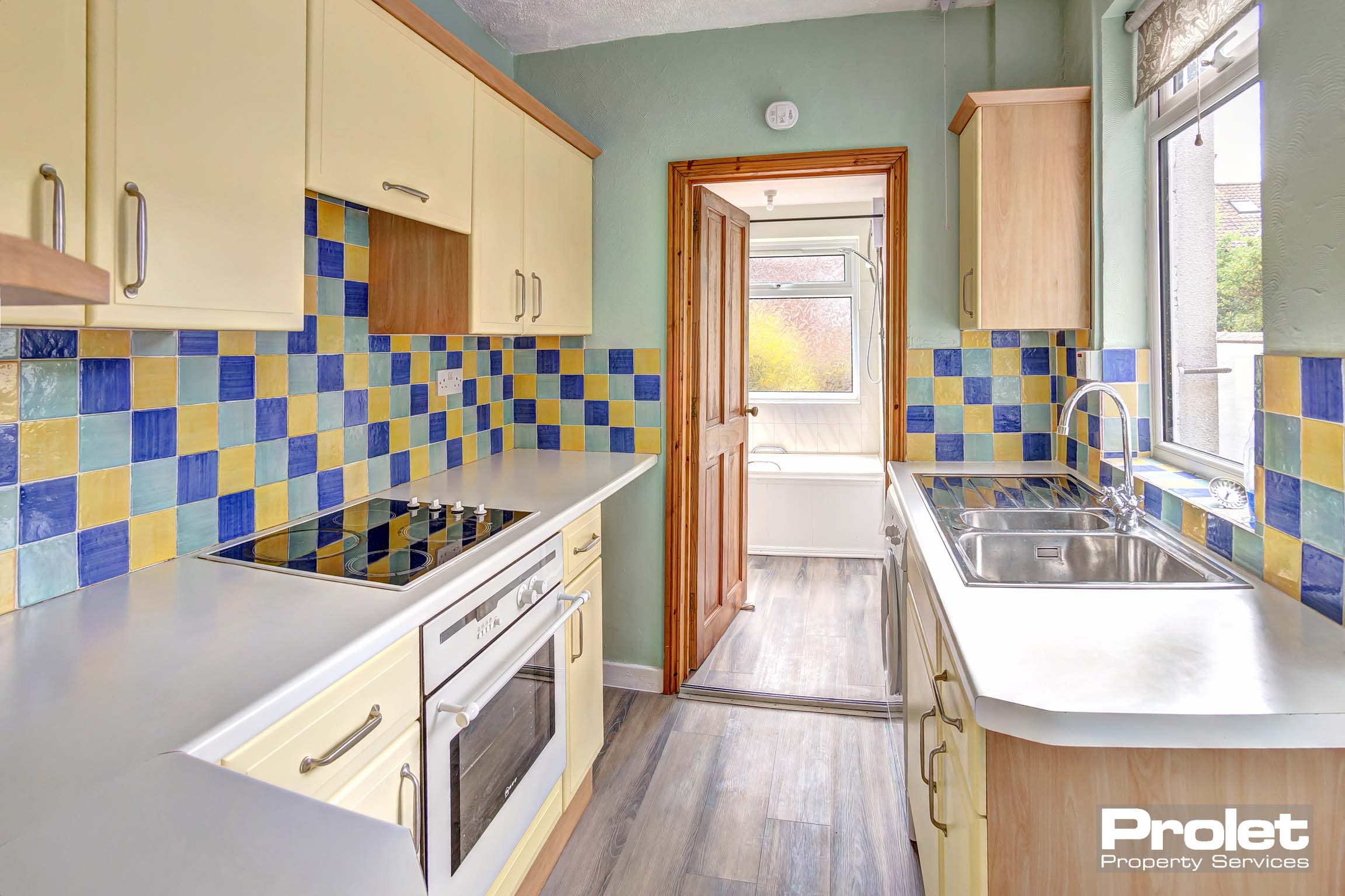 Kitchen leading to bathroom with vinyl flooring. Buttermilk coloured kitchen with white work tops. Walls painted green and yellow, green and blue wall tiles. Built in oven and hob. Gifted washing machine