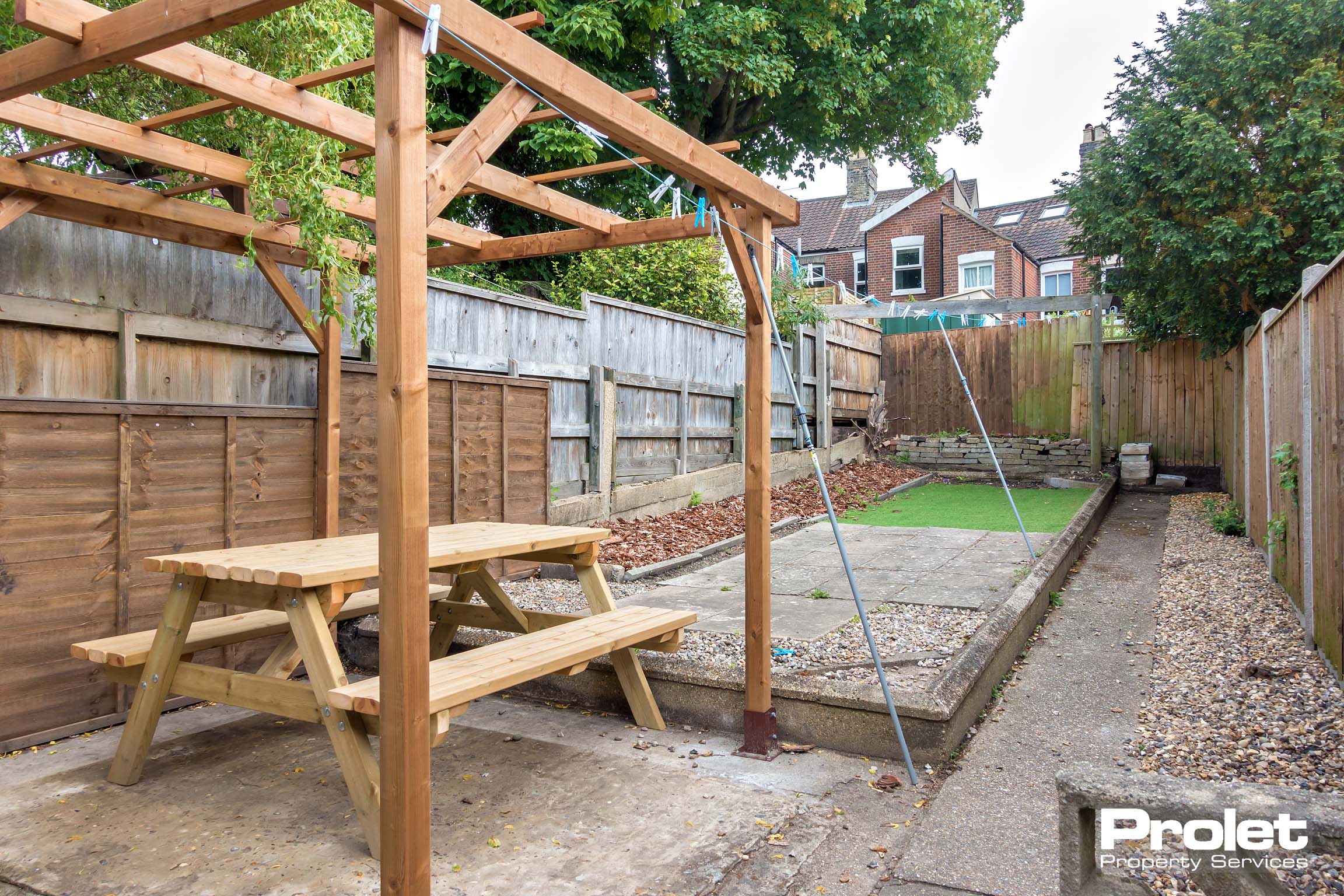 Garden area with wooden canopy with a picnic bench underneath, a concrete area and a grass area.