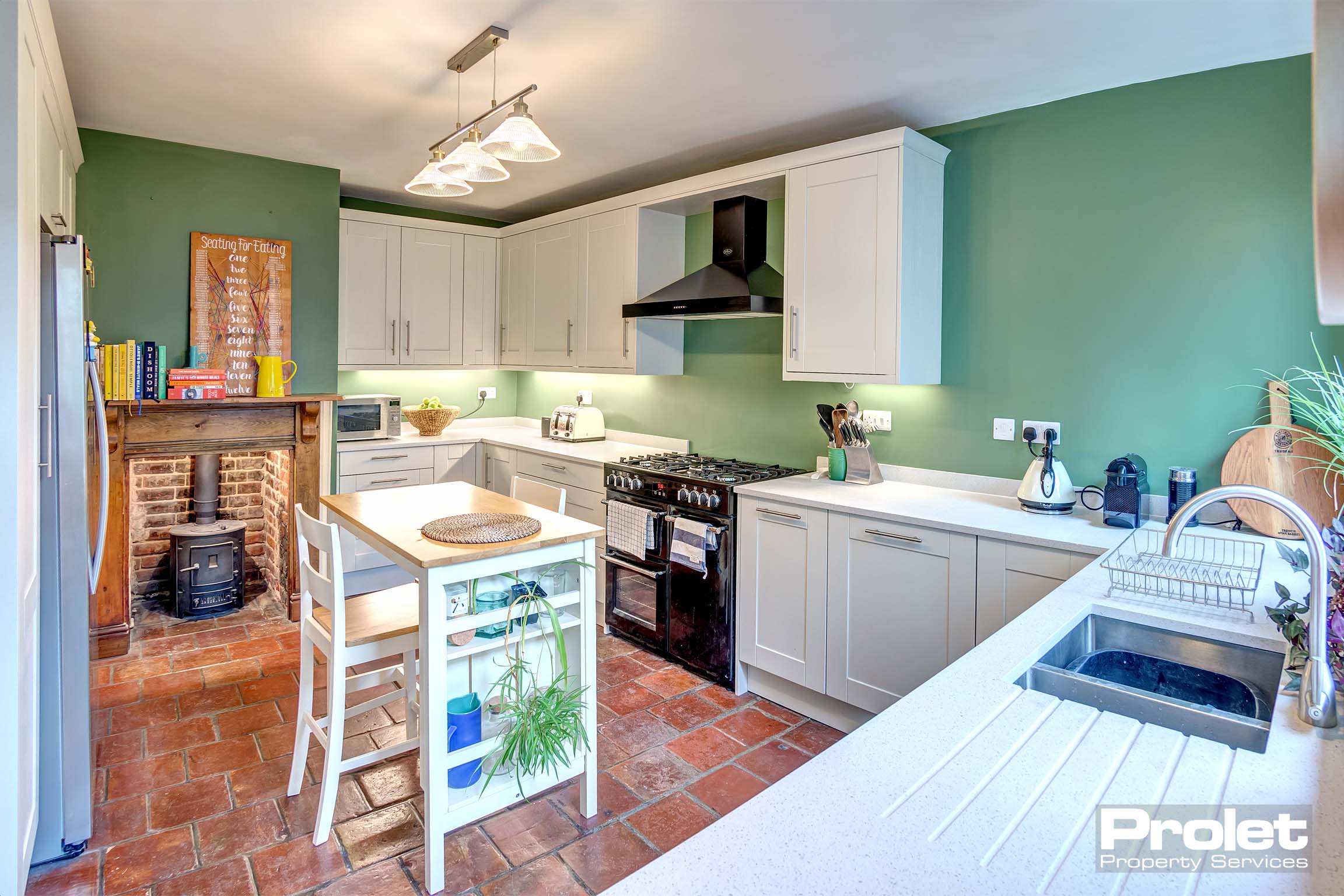 Modern kitchen with period features. Tiled floor, and wood stove in brick inglenook. White cabinets and worktop