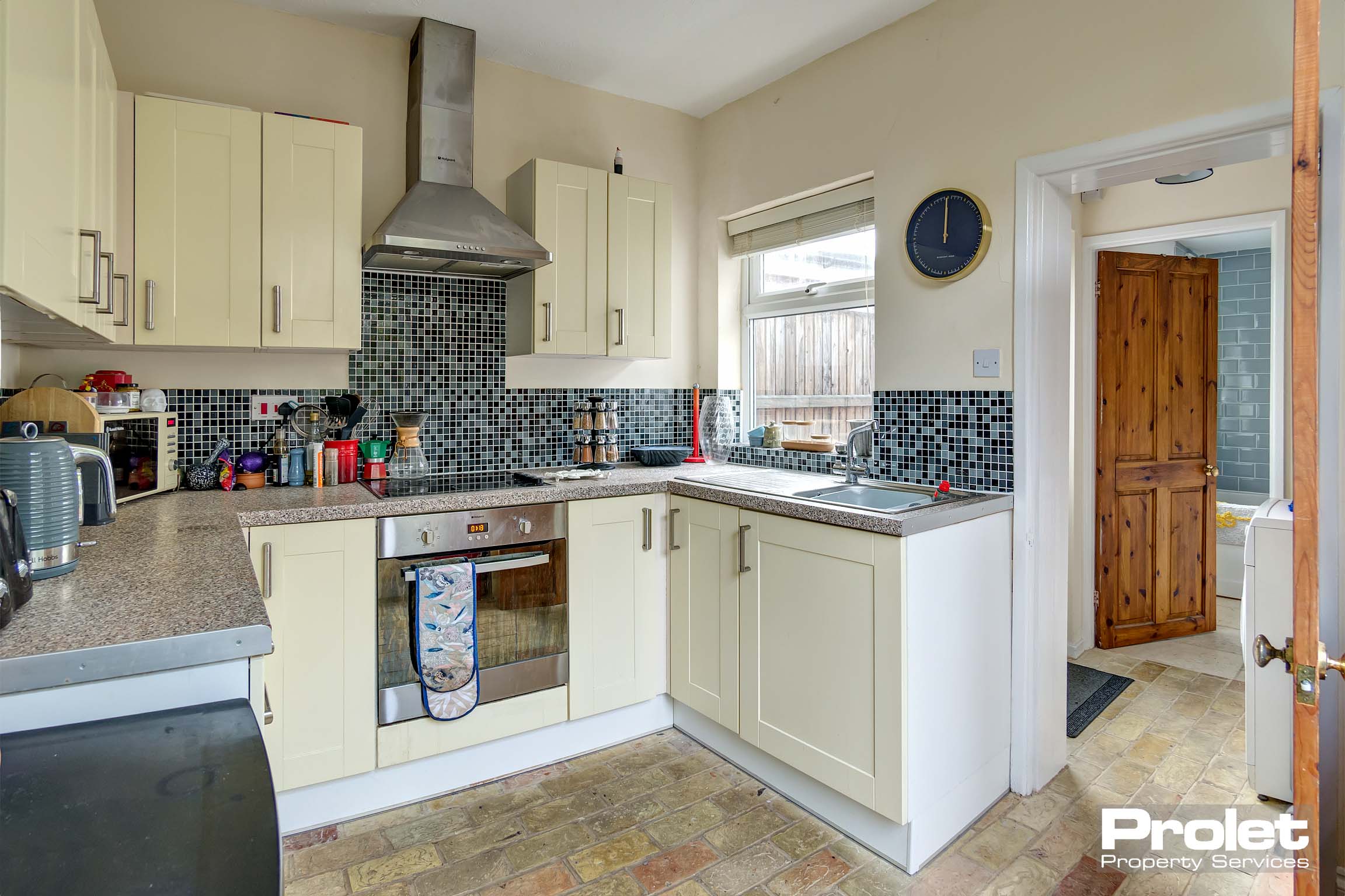Kitchen with brick floor, cream units, black worktops, cream paint and mosaic tiles to wall