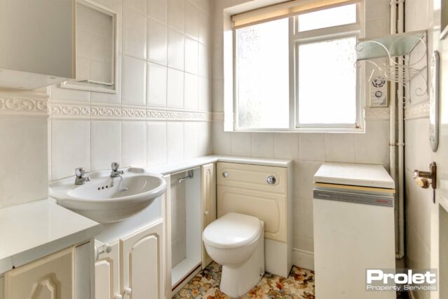 White tiled walls, sink and toilet with cream units. Floral carpet and shelves