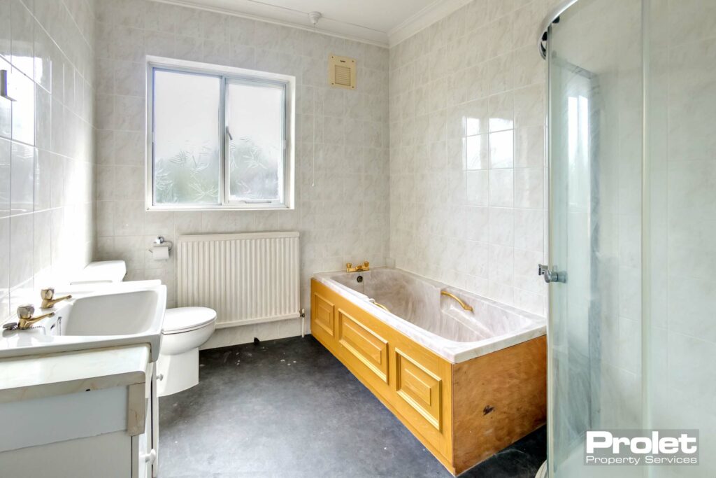 Walls tiles with grey flooring. Wooden bath panel with marbel effect bath. Separate cirner shower cubicle with white toilet and sink