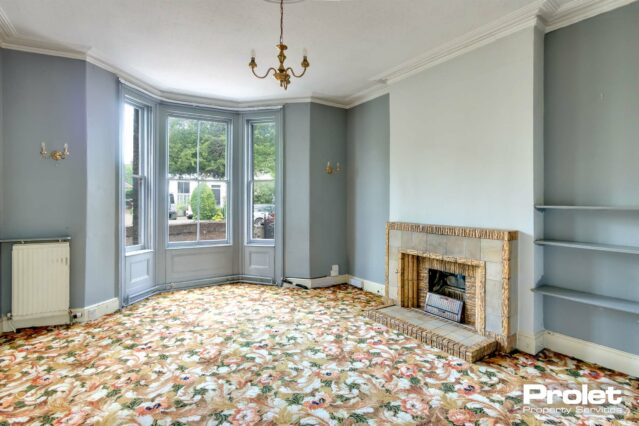 Bay window painted light grey with white walls, brick feature fireplace and floral carpet