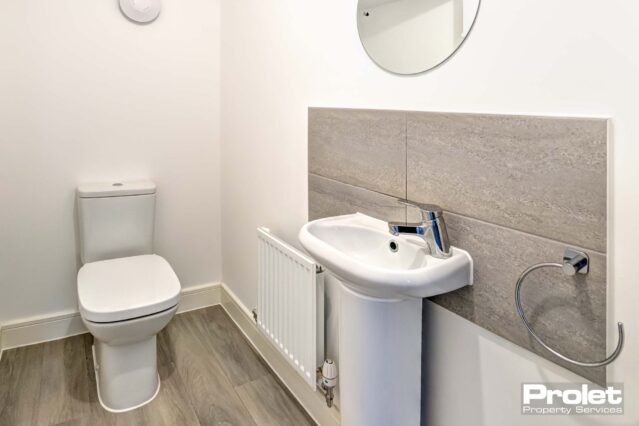 W.C with laminate flooring, white suite with white walls and tiled splash back. Mirror attached to wall