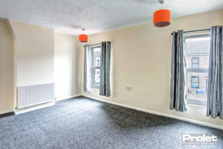Master bedroom with grey carpet and magnolia walls. Two orange lampshades and grey curtains lining two large windows.