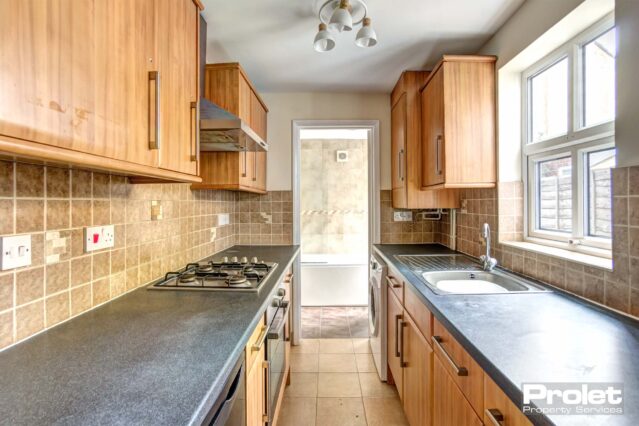 Beech effect worktops, grey worktops with brown wall tiles and flooring. Cooker, under counter fridge and washing machine.